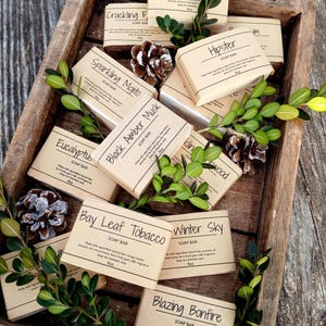 May include: A wooden tray filled with various handmade soap bars wrapped in brown paper. The soap bars have names like "Sparkling Mojito", "Black Amber Musk", "Bay Leaf Tobacco", and "Blazing Bonfire". Each bar is labeled with "SOAP BAR" and the weight in ounces.