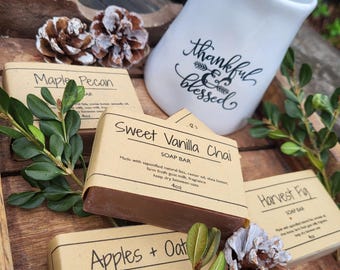 May include: A white ceramic pitcher with the text thankful & blessed, surrounded by several rectangular soap bars wrapped in brown paper labels. The labels feature names like Maple Pecan, Sweet Vanilla Chai, Harvest Fig, and Apples + Oat.