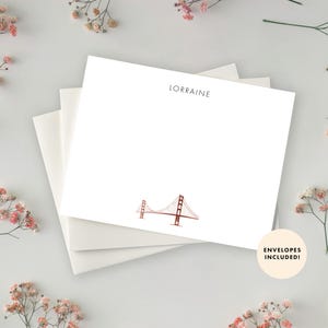 May include: White stationery set with the name "Lorraine" printed at the top. The stationery features a simple line drawing of the Golden Gate Bridge in red and brown. The text "Envelopes Included!" is printed on a circle in the bottom right corner.