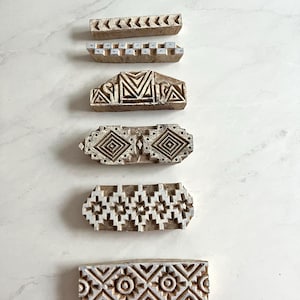 May include: Six wooden printing blocks with carved geometric patterns. The blocks are rectangular, with designs including chevrons, squares, and floral motifs. The wood has a natural, aged appearance, with visible grain and slight colour variations. The blocks are for textile printing.