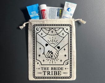 May include: A white canvas bag with a black design of two hands holding each other. The design is surrounded by stars and a crescent moon. The text "The Bride Tribe" is printed below the design.