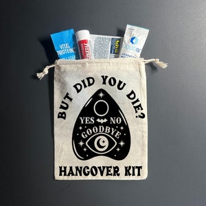 May include: A white canvas bag with a black design of a Ouija board with the text "But did you die?" and "Hangover Kit". The bag contains various items, including a blue packet of Vital Proteins Collagen, a blue packet of Liquid IV Hydration Multiplier, a white tube of Tylenol, and a white packet of Advil.