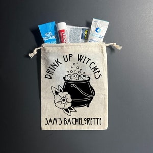 May include: A white canvas bag with a black cauldron and the text "Drink Up Witches Sam's Bachelorette" printed on it. The bag is filled with various small items, including a blue and white packet, a red and white tube, and a white packet with blue text.