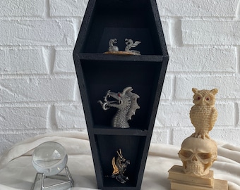 May include: Black coffin-shaped shelf with three shelves, each displaying a small silver dragon figurine. The shelf is on a white surface with a crystal ball and a skull with an owl on top.