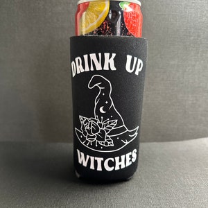 Peut inclure: Glacière noire pour canette avec du texte blanc qui dit "Drink Up Witches" et un chapeau de sorcière avec un croissant de lune et des étoiles.