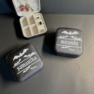 Puede incluir: Dos cajas de joyería negras con siluetas de murciélagos y nombres personalizados. Las cajas están cerradas y tienen un fondo blanco con los nombres "Samantha" y "Miranda" impresos en blanco.
