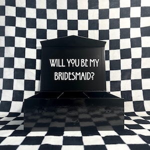 May include: Black box with a white lid that says "Will you be my bridesmaid?" The box is open and has four compartments inside.
