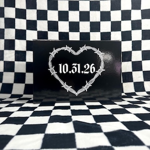Puede incluir: Caja negra con un diseño de corazón de alambre de púas blanco y la fecha 10.31.26 dentro del corazón. La caja está sobre una superficie a cuadros en blanco y negro.