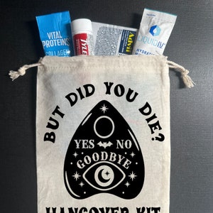 May include: A white canvas bag with a black design of a Ouija board with the text "But did you die?" and "Hangover Kit". The bag contains various items, including a blue packet of Vital Proteins Collagen, a red tube of Tylenol, a white packet of Advil, and a blue packet of Liquid IV.
