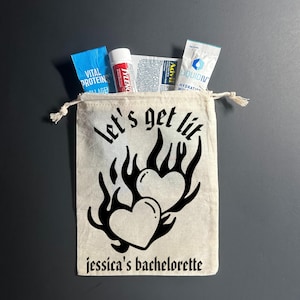 May include: A beige drawstring bag with the text "Let's get lit" and two heart-shaped flames. The bag contains small packets of Vital Proteins collagen, Tylenol, Advil, and Liquid I.V. The text "jessica's bachelorette" is printed below the design.