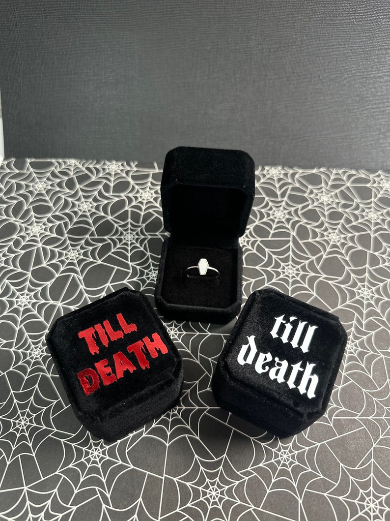 K&ouml;nnte beinhalten: Zwei schwarze Samt-Ringboxen mit den Worten "till death" in roter und wei&szlig;er Schrift. Die Box auf der linken Seite hat die Worte "till death" in roter Schrift. Die Box auf der rechten Seite hat die Worte "till death" in wei&szlig;er Schrift. Ein silberner Ring befindet sich in der ge&ouml;ffneten Box in der Mitte.