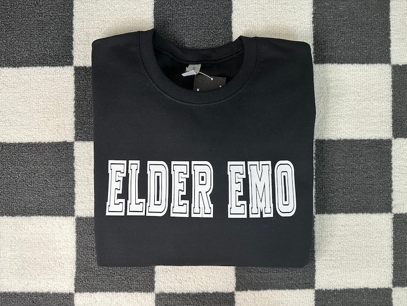 Puede incluir: Sudadera negra con texto blanco que dice "ELDER EMO" en una fuente de estilo universitario.