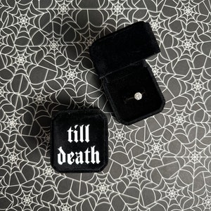 K&ouml;nnte beinhalten: Schwarze Samt-Ringbox mit den Worten "till death" in wei&szlig;en gotischen Buchstaben. Die Box ist ge&ouml;ffnet und ein Diamantring befindet sich darin.