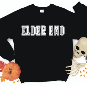 Puede incluir: Sudadera negra con texto blanco que dice "ELDER EMO". La sudadera est&aacute; rodeada de decoraciones de oto&ntilde;o, incluyendo una calabaza, hojas y ma&iacute;z dulce.