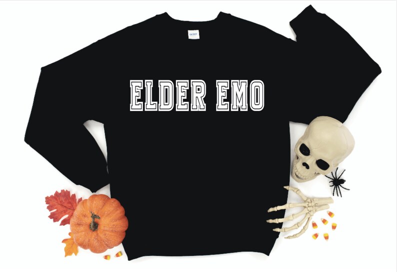 Puede incluir: Sudadera negra con texto blanco que dice "ELDER EMO". La sudadera est&aacute; rodeada de decoraciones de oto&ntilde;o, incluyendo una calabaza, hojas y ma&iacute;z dulce.