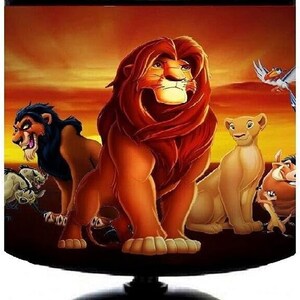 Pode incluir: Candeeiro de mesa preto com abajur com personagens de O Rei Leão. O abajur apresenta Simba, Scar, Nala e outros personagens contra um fundo de pôr do sol. A base da lâmpada é uma esfera preta.
