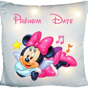 Peut inclure: Un oreiller blanc avec un motif Minnie Mouse rose. Minnie Mouse porte un nœud rose et a des ailes d'ange. L'oreiller porte les mots "PRENOM DATE" imprimés dessus.