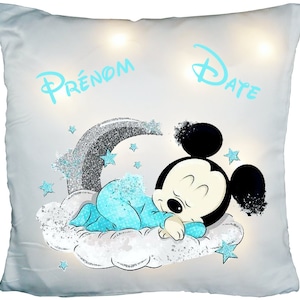 Peut inclure: Un coussin carré gris clair avec un motif de Mickey Mouse endormi. Mickey porte un body bleu et repose sur un nuage. Le coussin porte les mots "Prénom" et "Date" en bleu clair, ainsi que des étoiles et un arc-en-ciel.