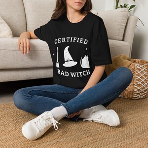 Può includere: Una t-shirt nera a maniche corte con una stampa grafica bianca che include una scopa, un cappello da strega e la sagoma di un gatto, con il testo "CERTIFIED BAD WITCH" stampato in un carattere bianco e grassetto sul petto.