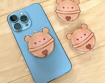 May include: Three brown bear-shaped phone charms with a bell design. The charms are made of clear plastic with a brown bear design and a heart-shaped bell. The charms are on a light wood surface.