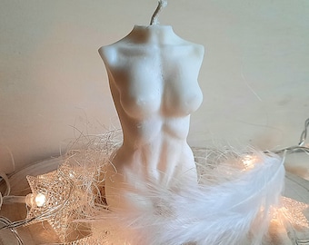 May include: A white candle shaped like a human torso. The candle is sitting on a wooden surface with white feathers and string lights.