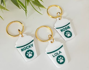 Peut inclure: Trois porte-clés en forme de tasse à café blanche avec des anneaux dorés. Chaque porte-clés a une empreinte de patte verte et un nom : "Lola", "Rocky" et "Rosie".