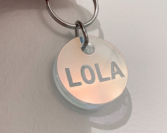 May include: A round, iridescent white pet tag with a silver key ring. The tag has the name "LOLA" printed in black letters.