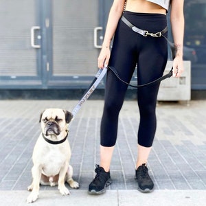 Op de afbeelding: Een persoon met zwarte leggings en een riem loopt met een lichtgrijze hondenriem met de naam "Huxley" erop gegraveerd. De hond is een kruising tussen een mops en zit op een grijs trottoir.