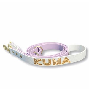 Op de afbeelding: Een witte leren hondenriem met een lavendelkleurige leren band en gouden beslag. De riem is gepersonaliseerd met de naam "KUMA" in gouden letters.