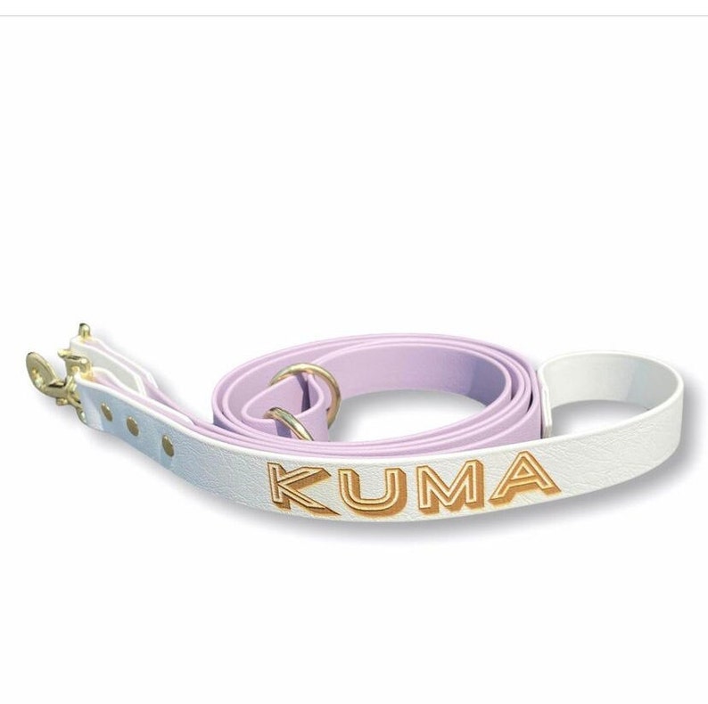 Op de afbeelding: Een witte leren hondenriem met een lavendelkleurige leren band en gouden beslag. De riem is gepersonaliseerd met de naam "KUMA" in gouden letters.