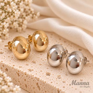 May include: Two pairs of stud earrings, one pair in a polished gold tone and one pair in a polished silver tone. Each earring features a smooth, rounded, dome-shaped design with a standard post backing. The text Manna Los Angeles is visible.