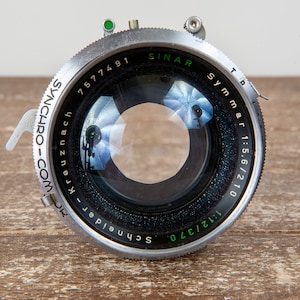 Op de afbeelding: Close-up van een zilver-zwarte cameralens met de tekst "Schneider-Kreuznach" en "Synchro-Com" op de buitenste ring. De lens heeft een helder glazen midden en staat tegen een houten achtergrond.