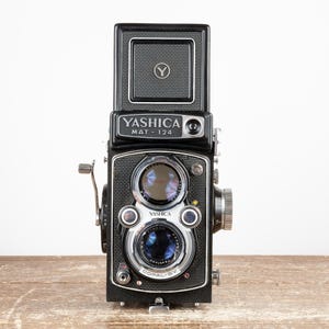May include: A black Yashica Mat-124 twin-lens reflex camera. The camera has a square viewfinder, two lenses, and various dials and levers. The name "Yashica Mat-124" is printed on the front. The camera is on a wooden surface.