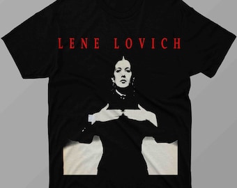 May include: Black t-shirt featuring a red text that reads "LENE LOVICH" above a black and white image of a person. The person is wearing a dark top and has their hands crossed over their chest.