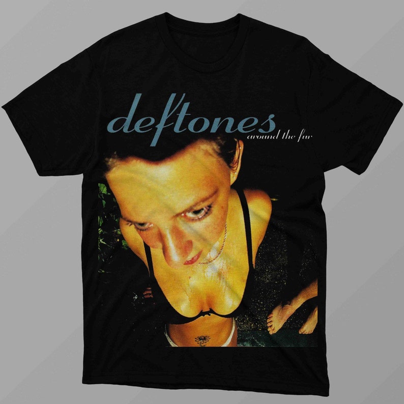 May include: Black t-shirt featuring the band name "Deftones" in teal script above the words "around the fur". The shirt has a graphic of a person looking down. The person is wearing a black top.
