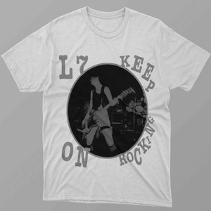 May include: A white t-shirt with a black and white image of a person playing guitar. The text "L7 KEEP ON ROCKING" is printed around the image.