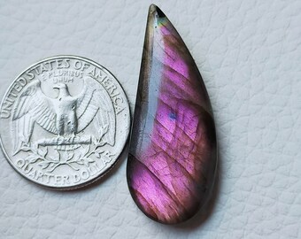 May include: A teardrop-shaped, polished labradorite gemstone with vibrant purple and pink hues. The gemstone is next to a US quarter for size comparison. The stone has a dark gray border and a pointed top.