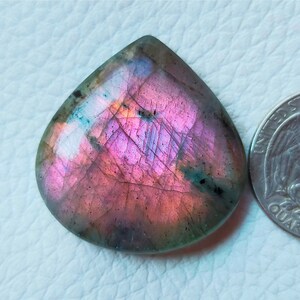 May include: A pear-shaped, polished labradorite gemstone with a vibrant display of pink, purple, and teal iridescence. The stone is next to a quarter for size comparison, showcasing its unique color play and natural beauty.
