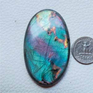 May include: Oval-shaped labradorite gemstone with iridescent blue, purple, and gold hues. The polished stone displays natural patterns and is next to a quarter coin for size comparison. The gemstone is a beautiful example of natural mineral artistry.