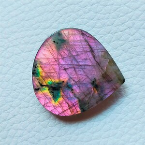 May include: A teardrop-shaped gemstone with a vibrant display of colors. The stone features a blend of purple, pink, and hints of green and yellow. The surface has a textured appearance with visible lines and patterns, creating a unique and eye-catching aesthetic.