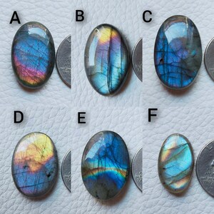 May include: Six oval-shaped labradorite gemstones with iridescent flashes of blue, purple, and yellow. Each stone is displayed next to a quarter for size comparison. The gemstones have a polished, smooth surface.