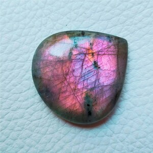 May include: A polished, teardrop-shaped labradorite gemstone. The stone displays a vibrant play of color, with iridescent flashes of pink, purple, and blue across its surface. The gemstone is a natural, organic shape, and is likely used for jewelry or display.
