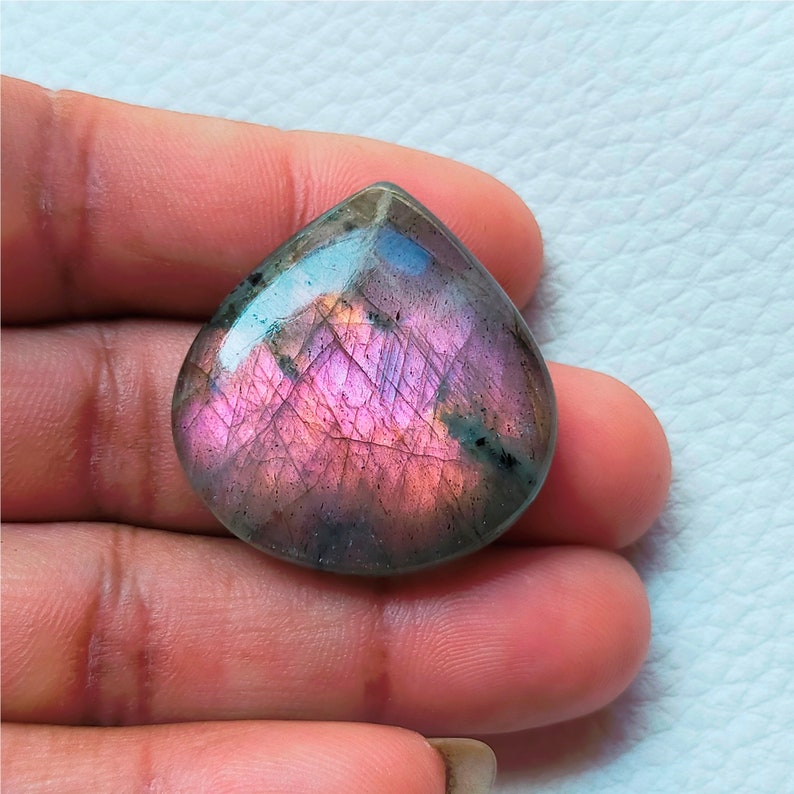 May include: A pear-shaped, polished gemstone with a vibrant display of colors. The stone features a mix of pink, purple, and blue hues, with visible internal patterns. The gemstone is held in a hand, showcasing its size and shape.