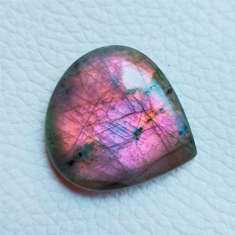 May include: A polished, teardrop-shaped labradorite gemstone. The stone displays a vibrant play of color, with iridescent flashes of pink, purple, and blue across its surface. The gemstone is set against a white background.