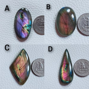 May include: Four polished labradorite gemstones in different shapes: oval, rounded oval, kite, and teardrop. Each stone displays iridescent flashes of blue, purple, and gold. A U.S. quarter coin is next to each stone for size comparison. The gemstones are on a white surface.