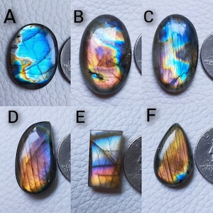 May include: Six polished labradorite gemstones in various shapes: oval, teardrop, and rectangular. The stones display iridescent flashes of blue, purple, and gold. Each stone is next to a quarter for size comparison. The gemstones are set against a light background.