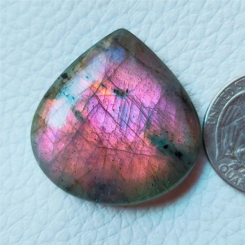 May include: A pear-shaped, polished labradorite gemstone with a vibrant display of pink, purple, and teal iridescence. The stone is next to a quarter for size comparison, showcasing its unique color play and natural beauty.