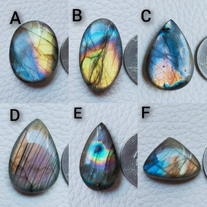 May include: Six polished labradorite gemstones in various shapes: oval, teardrop, and triangular. The stones display iridescent flashes of blue, gold, and purple. Each stone is next to a quarter for size comparison. These gemstones are ideal for jewelry making.