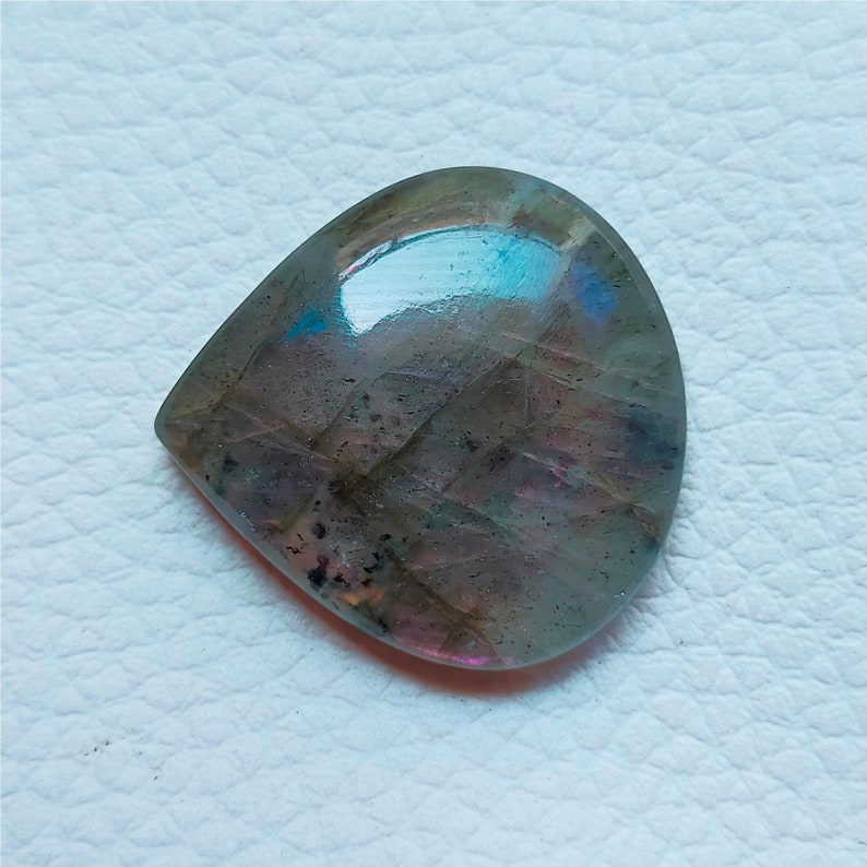 May include: A polished, teardrop-shaped labradorite gemstone. The stone displays a mix of gray, purple, and blue hues with iridescent flashes. The surface is smooth and reflective, showcasing natural patterns.