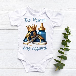May include: White baby onesie with the text "The Prince has arrived" in blue lettering. The onesie features a gold crown, blue baby shoes, and a blue baby bottle.